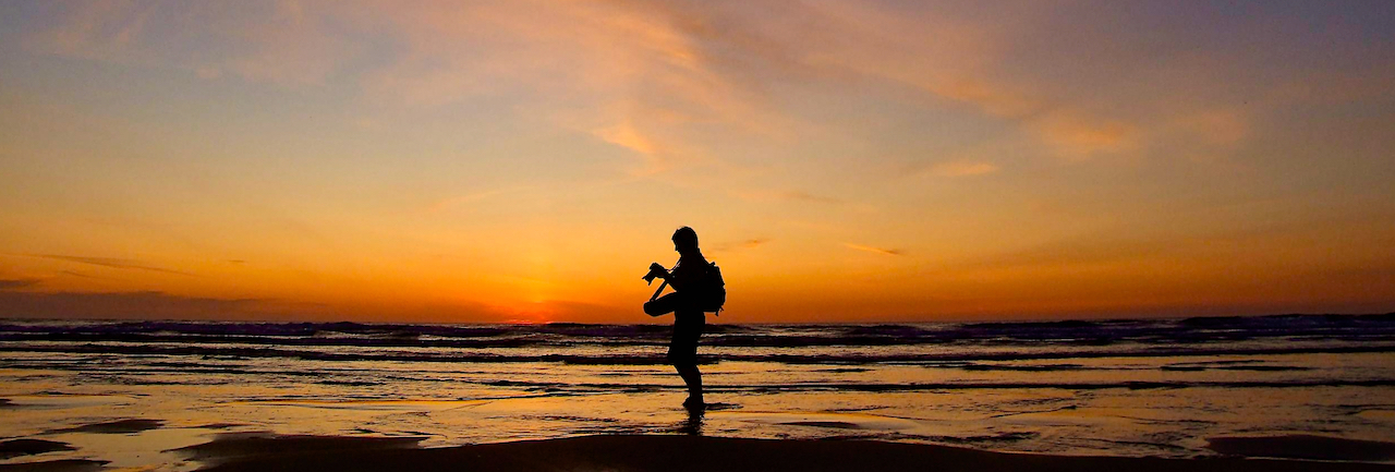 A photograph of Emma Cox silhouetted against the sunset with her camera and tripod. She is on a beach, standing in the shallows with the sea behind her. The sunset is a deep line of red on the horizon working through hues of orange, yellow and dusky pinks higher up.