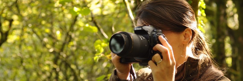 A woman holding a Nikon DSLR to her eye in a woodland setting