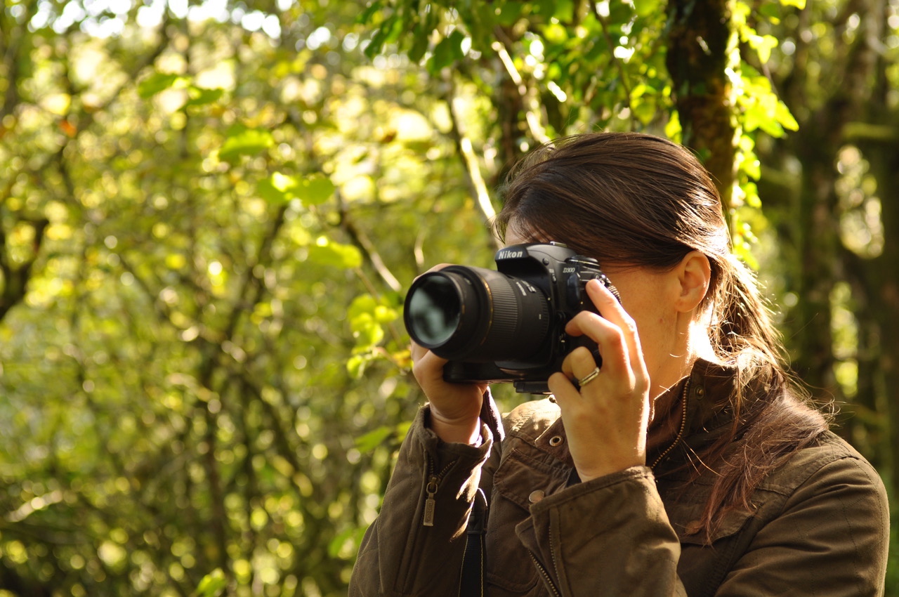 A woman holding a Nikon DSLR to her eye in a woodland setting
