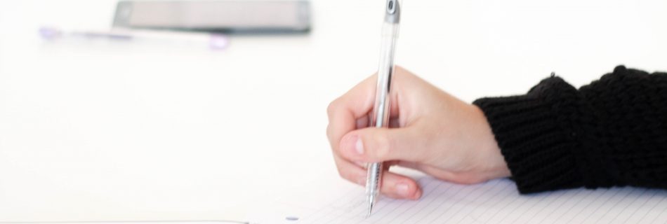 Photograph of a right hand holding a pen over a note book beside a laptop. There are words written on the wall in the background, "Life is a Journey, enjoy it."