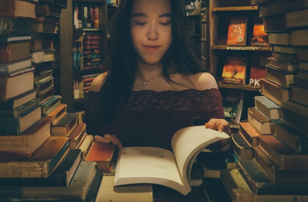 Books stacked so there is a circular window opening where a woman with dark hair is reading a book through the aperture.