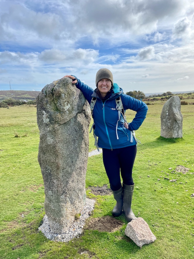 A photo of a stone from the Hurlers Stone Circle with Emma Cox posing next to it in a blue coat.