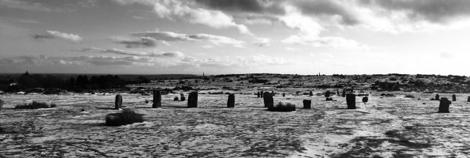 A black and white photograph of the Hurlers Stone Circles in Cornwall. There is snow on the ground and the stones are silhouetted against the white sky.