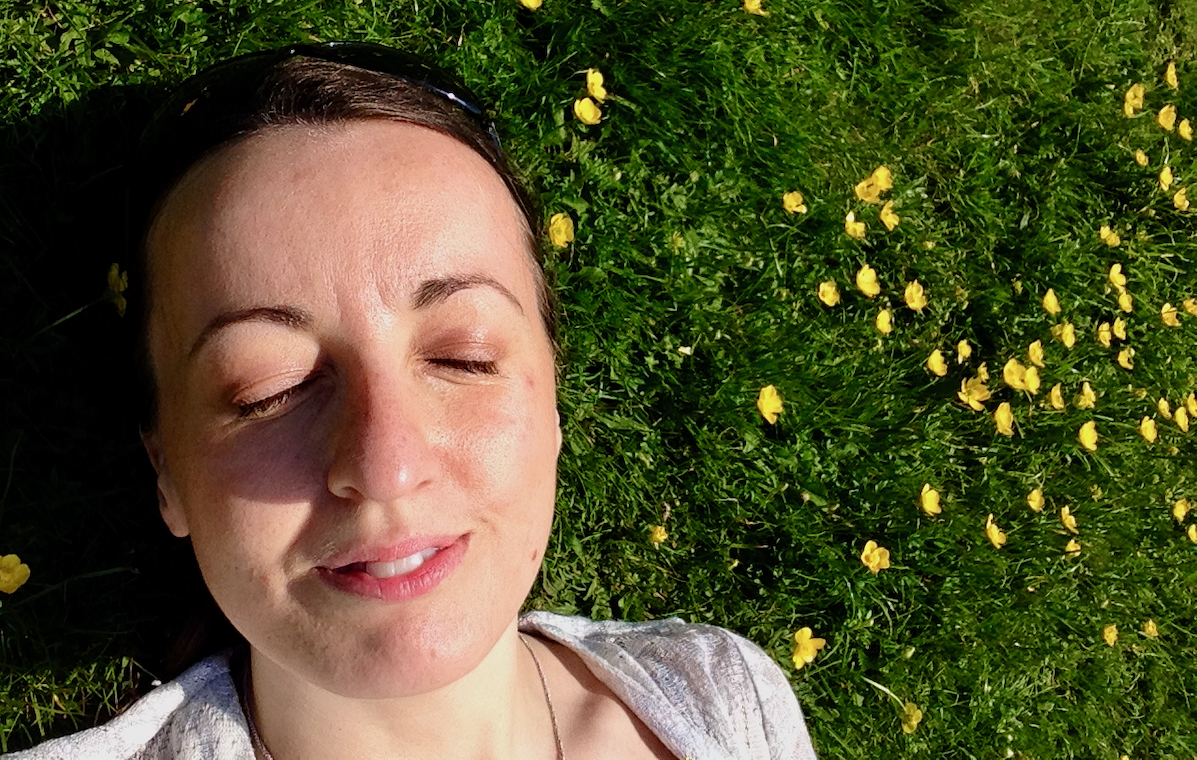 A portrait photograph of a woman with dark hair lying in grass dotted with yellow buttercups.