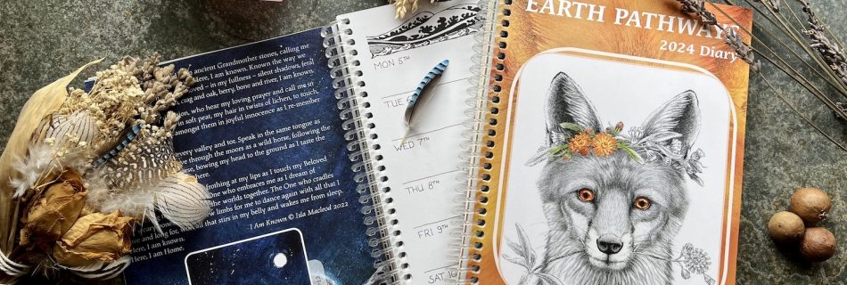 Two copies of the 2024 Earth Pathways Diary showing the front cover with an illustration of a fox and an open page with the diary layout and a painting of Mulfra Quoit by Emma Cox. The diaries are on a slate background surrounded by dried flowers, a pink quartz crystal, gall oaks and a wooden wand.