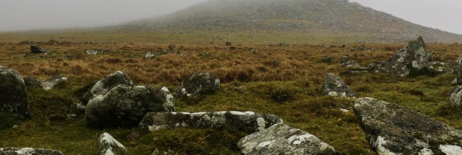 Moorland landscape with granite rocks in the foreground and a granite tor shrouded by mist in the background.