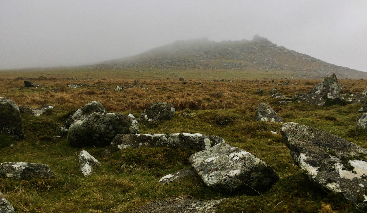 Moorland landscape with granite rocks in the foreground and a granite tor shrouded by mist in the background.