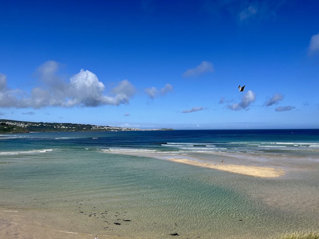 A sandy beach with azure to dark blue sea with blue skies and light cloud. A headland is in the distance.