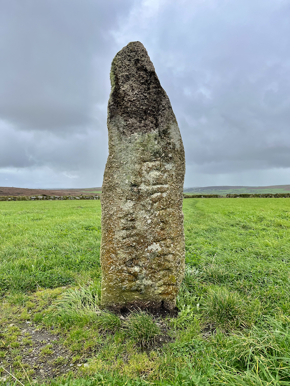 A standing stone with Latinised Cornish text. It stands in a field on a cloudy day.