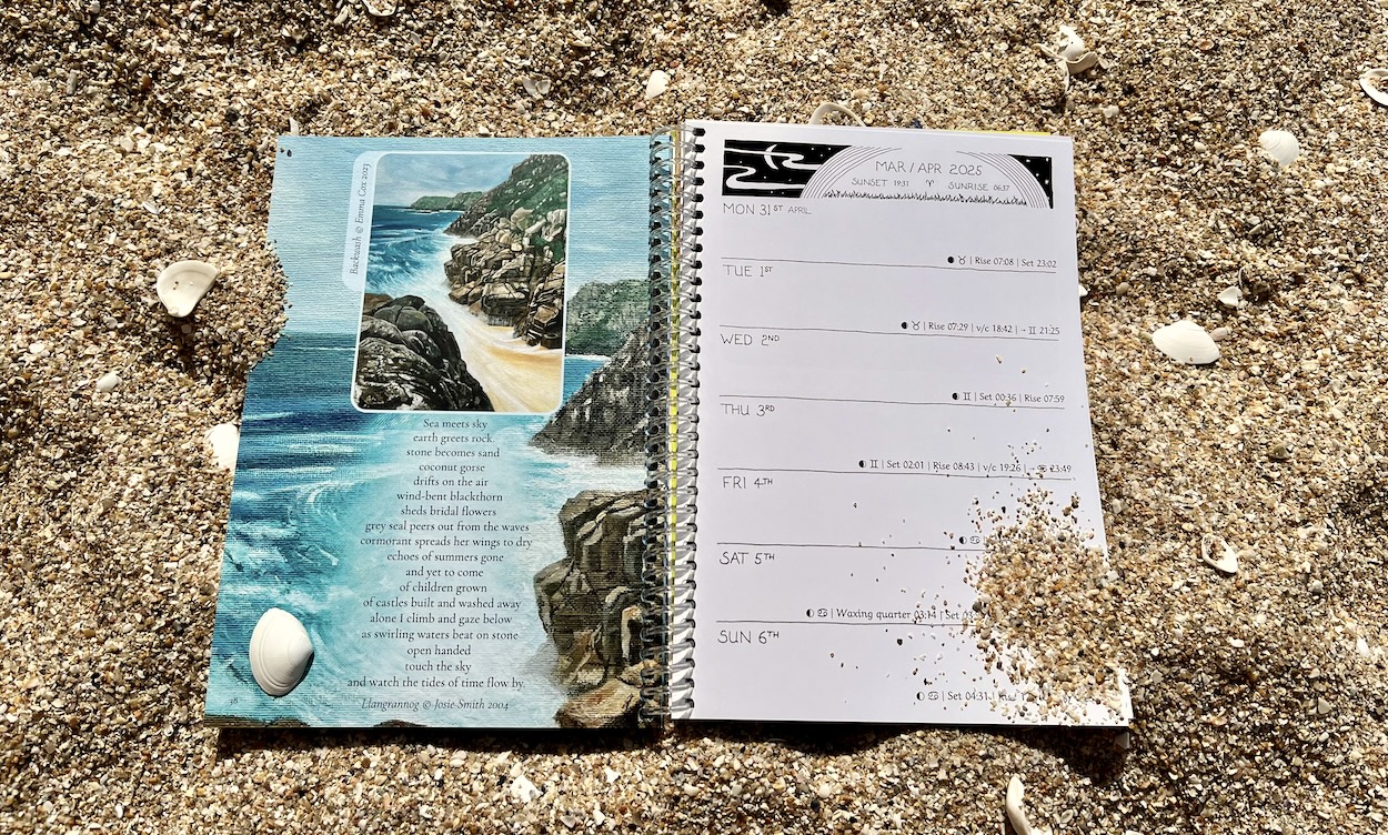 a photograph of the 2025 earth pathways diary lying on the beach open to a week in December depicting a photography in black and white of a standing stone accompanied by text.