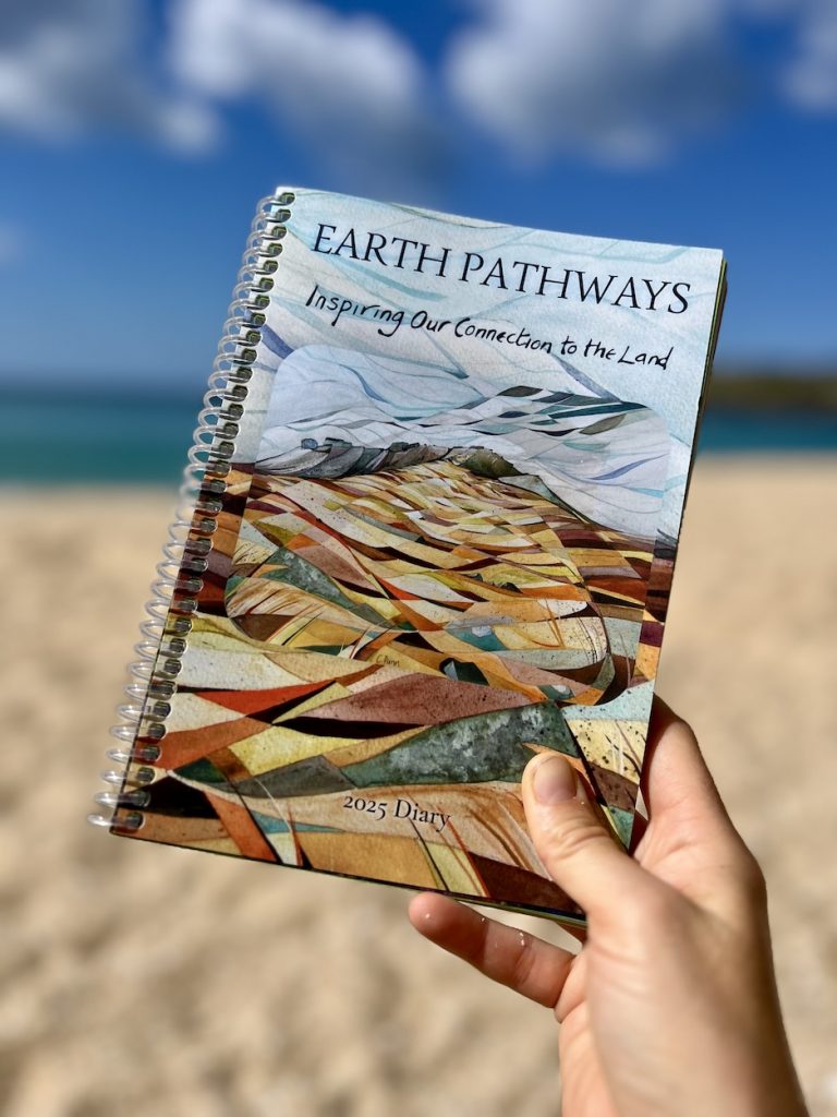 A photograph of a woman's right hand holding the Earth Pathways 2025 diary against a blurred background of the sea and sand. The diary depicts an artists painting of a blustery hill top.
