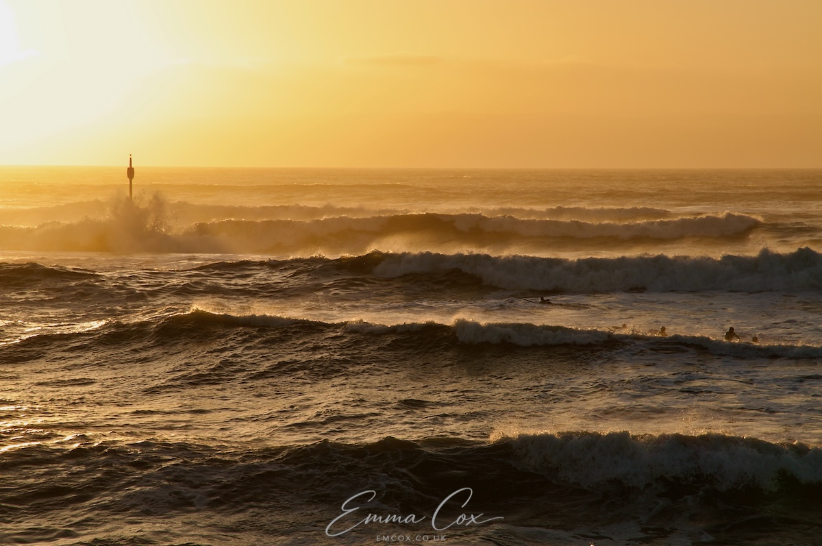 A photograph from the cliff top looking across Bude bay. It is the golden hour near sunset. Large sets of waves are populated by surfers. In the middle left of the image is a pole with a barrel on the top - a marker for the boats coming inland - which has been hit by a breaking wave.