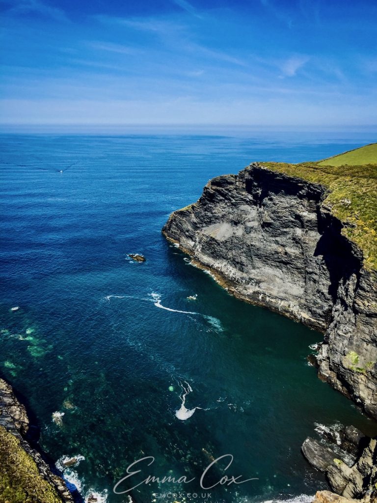 A photograph viewed from the Cornish clifftop looking out to sea. It is a calm sea and a blue sky. The small cove is surrounded by high, dark cliffs banded with white quartz.
