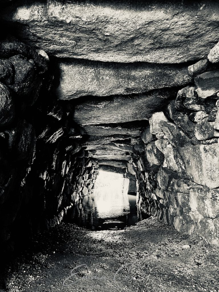A black and white photograph of a Cornish Fogou. It shows the inside of this manmade ancient underground drystone enclosure. The passage is lined with granite stone walls with a large lintel ceiling. The floor is soil and half flooded by rainwater.