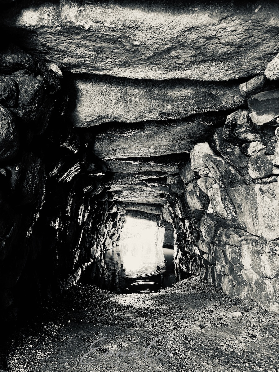 A black and white photograph of a Cornish Fogou. It shows the inside of this manmade ancient underground drystone enclosure. The passage is lined with granite stone walls with a large lintel ceiling. The floor is soil and half flooded by rainwater.