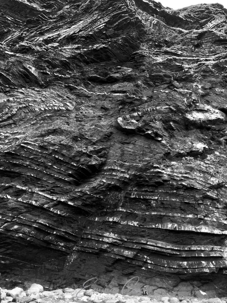A black and white photograph of a high cliff viewed from the beach. Geological bands of dark rock and white quartz make layers and patters. A tiny figure sat on the beach gives a sense of the immense scale of the cliff.