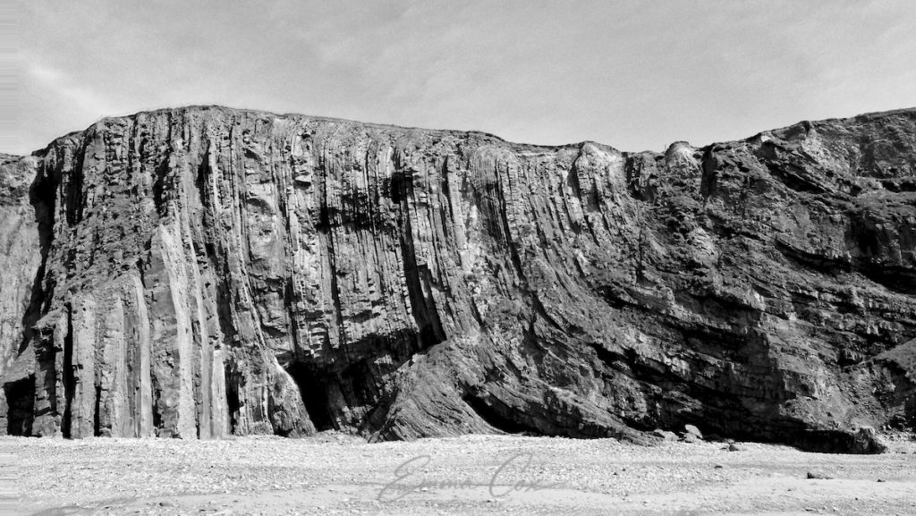 Black and white photograph of a cliff face showing the Cornish geology. Sediment layers in the rock, which would have been horizontal, are now vertical.