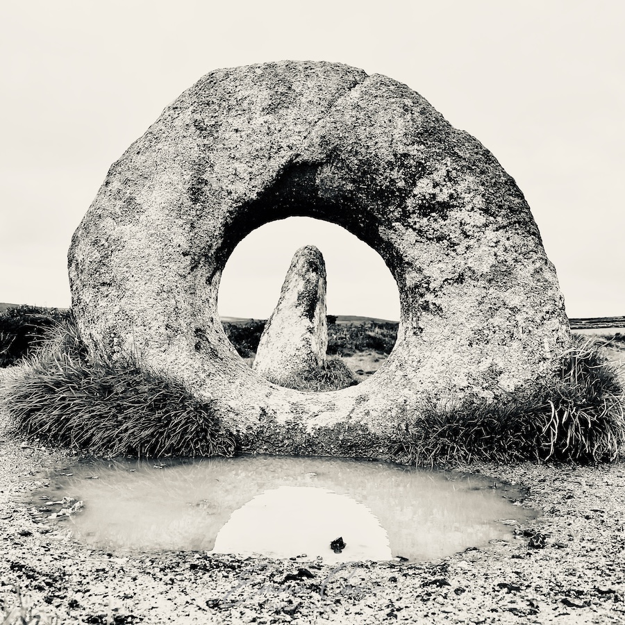 A black and white photograph of Men an Tol, a megalithic granite standing stone with a perfect hole in the centre. Framed within is another standing stone.
