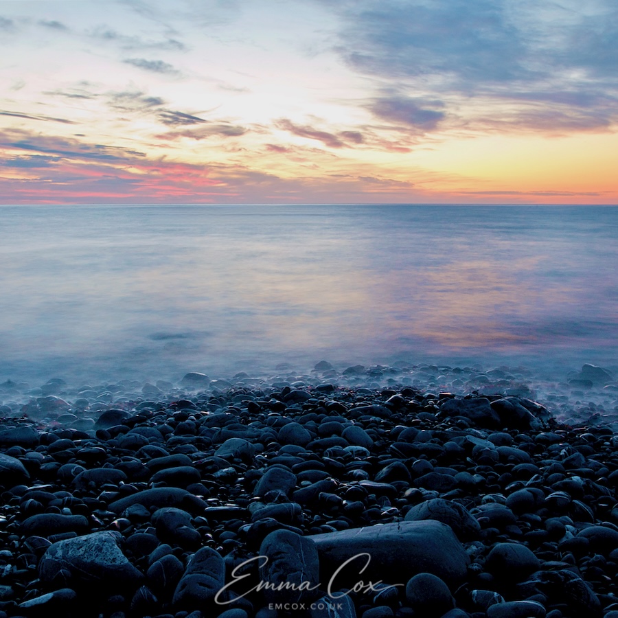 A long exposure photograph of a Cornish stony beach at sunset. The milky calm water curls mist-like across the stones. On the horizon, golden and pinky hues touch the sky.