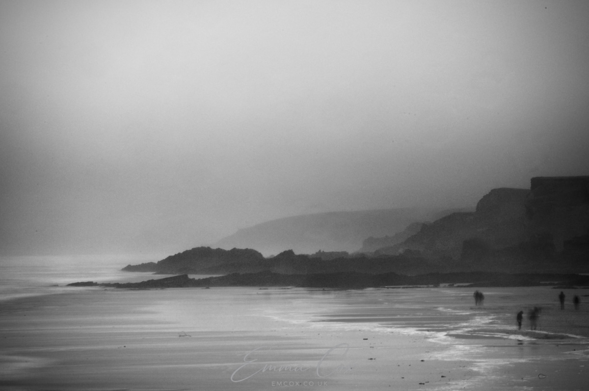 A black and white photograph viewing along the Cornish coastline. It is a misty day so the distant rocky headlands fade into the murk.