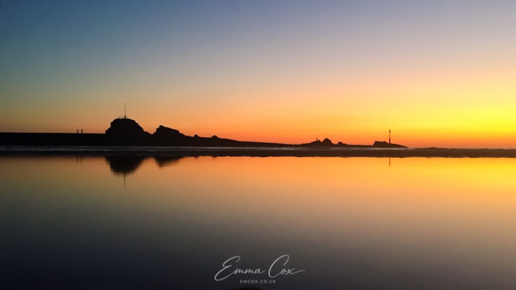 Bude's breakwater and the sunset are reflected in the mirror-like water.