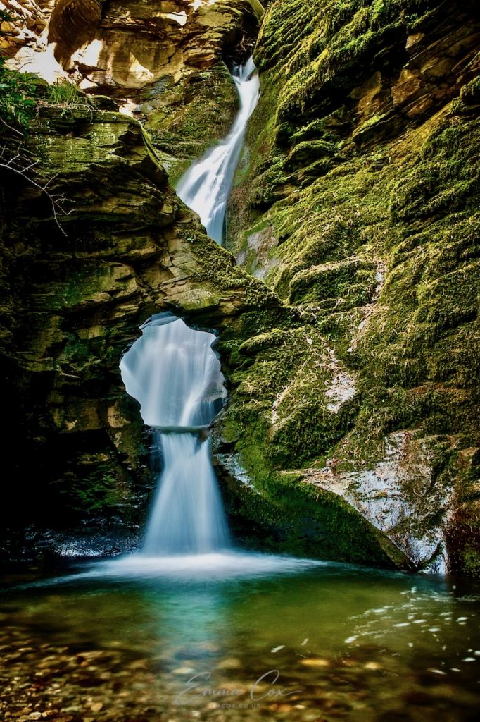 A long exposure photograph of a waterfall cascading down a mossy rock face. The water fall reaches a basin framed by an arch (kieve) and pours through it into the stream.