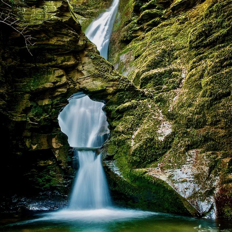 A long exposure photograph of a waterfall cascading down a mossy rock face. The water fall reaches a basin framed by an arch (kieve) and pours through it into the stream.