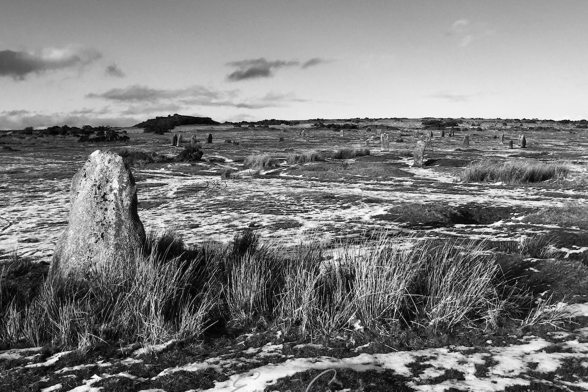 A black and white photograph of open moorland covered in a light dusting of snow. In the foreground is a stone from one of the three stone circles which makes the Hurlers. In the middle distance is the central stone circle.