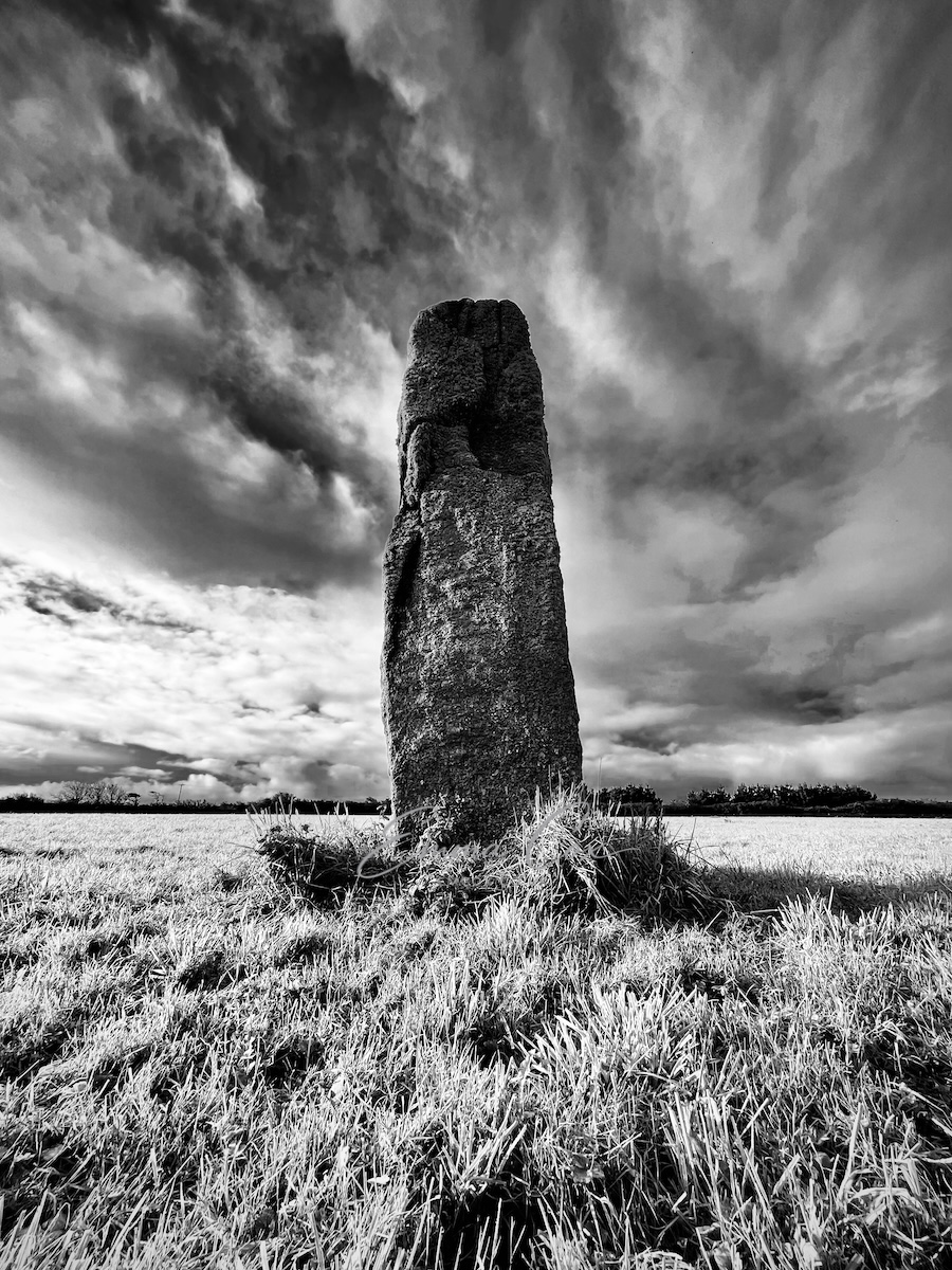 A black and white photograph of a tall granite Cornish standing stone in rough ground beneath a dramatic sky