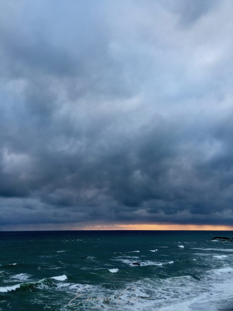 A photograph of a Cornish dark, stormy sea with blue/grey turbulent clouds overhead. At the horizon, there is a thin pinkish/orange hint of the sunset.