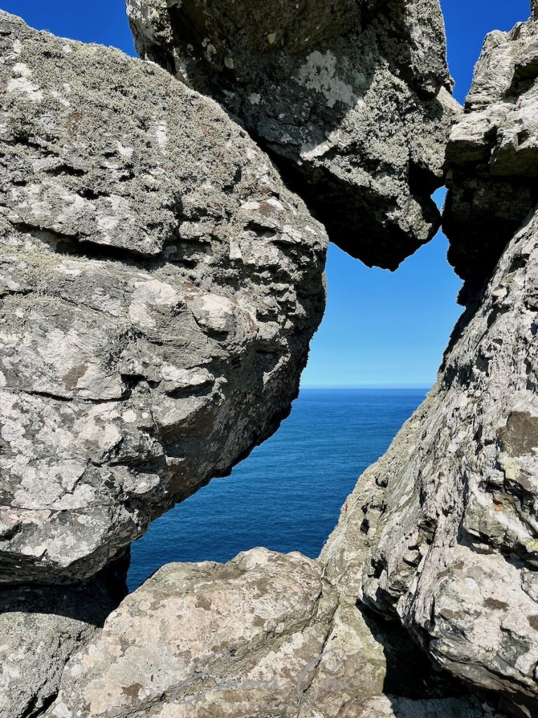 A photograph of a Cornish granite outcrop at Zennor headland. The boulders create a frame with a narrow window looking out to blue sea and sky.