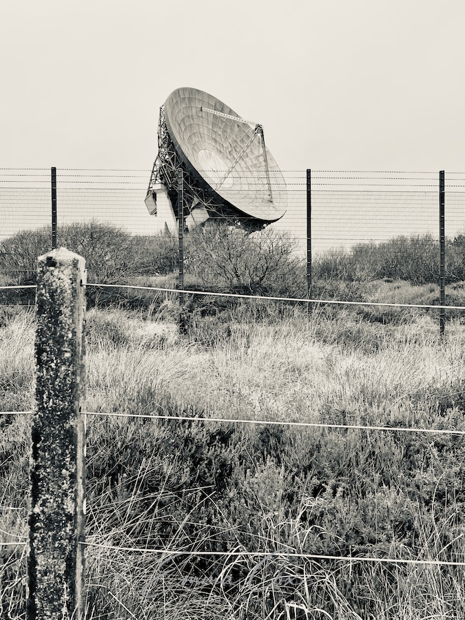 A black and white photograph of the Goonhilly Satellite Earth Station. A satellite dish points to the sky from behind a security fence. A livestock fence is in the foreground.