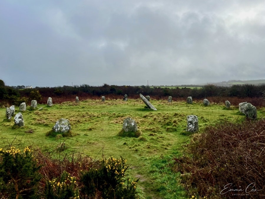 a stone circle on rough grassland surrounded by winter bracken and gorse. There is a central stone which leans to the left.